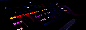 Due mani operano su una console di controllo Barco EC-210 in un ambiente buio. La console è ricca di pulsanti che si illuminano di vari colori, tra cui rosso, arancione, giallo, viola e bianco. Una luce illumina leggermente le mani, creando un contrasto nel buio