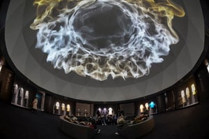 Una vista di una cupola. Sul soffitto della cupola è proiettata un'immagine astratta luminosa in tonalità di bianco e oro. Intorno alla sala, in sei sezioni numerate con didascalie in coreano, sono esposti abiti appesi. Diverse persone sono sedute su divanetti curvilinei o in piedi