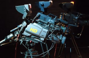 Tre videocamere professionali Sony, montate su treppiedi, sono disposte in un ambiente scuro. La videocamera in primo piano con la scritta 'HD' in giallo è ben visibile. Le altre due videocamere sono meno illuminate. Tutte le telecamere sono dotate di vari accessori, cavi e lenti