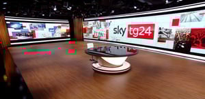 Studio TV di SKY TG24, due grandi videowall fanno da sfondo ad un tavolo rotondo con piano in materiale riflettente. A soffitto si nota un'importante schieramento di proiettori luce, mentre il pavimento è ricoperto di parquet